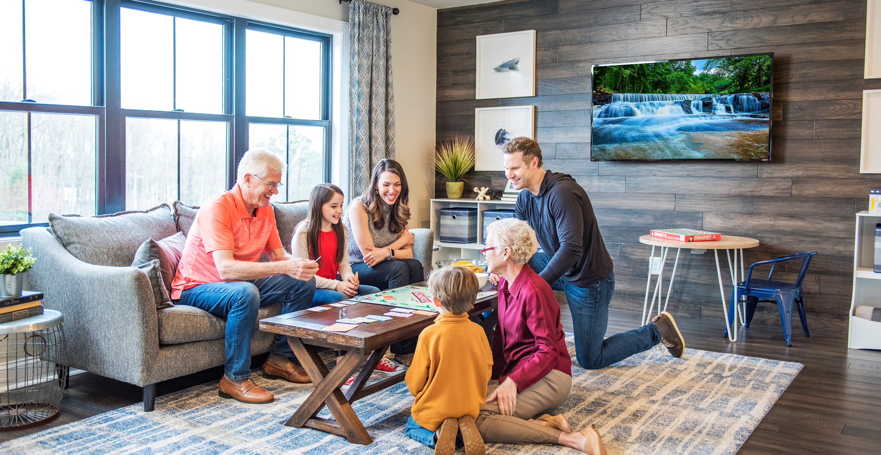 A multi-generational family gathers in a living room to play a board game together. The room is cozy with a TV and warm decor.