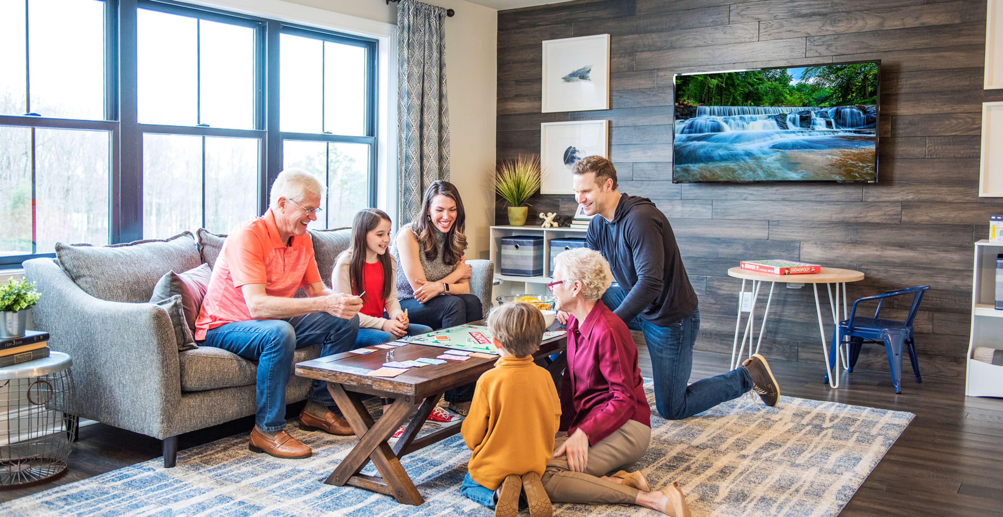 A multi-generational family gathers in a living room to play a board game together. The room is cozy with a TV and warm decor.