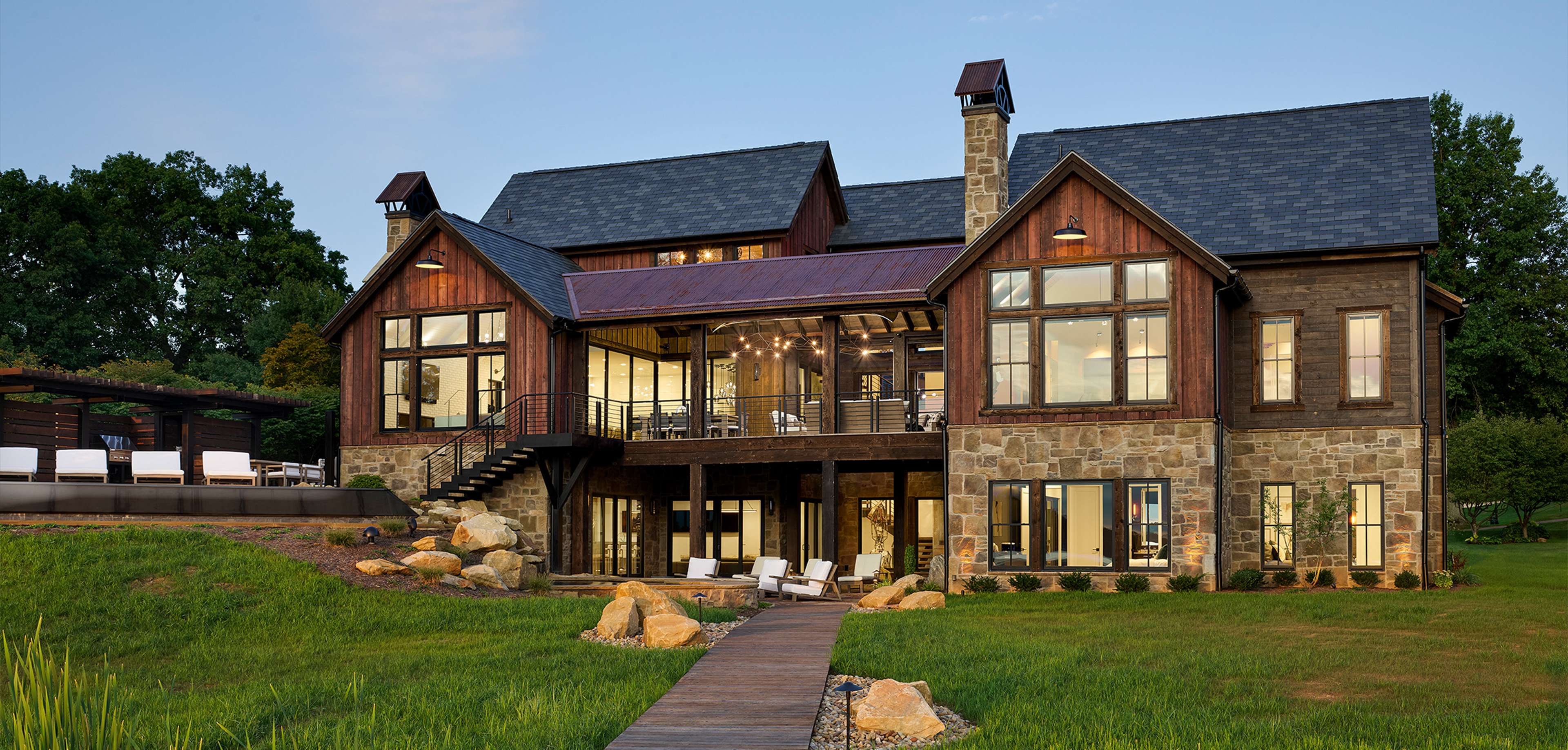 The exterior of a modern rustic home has a mix of stone and wood, large windows, and a cozy deck. The design blends natural materials with contemporary elements.