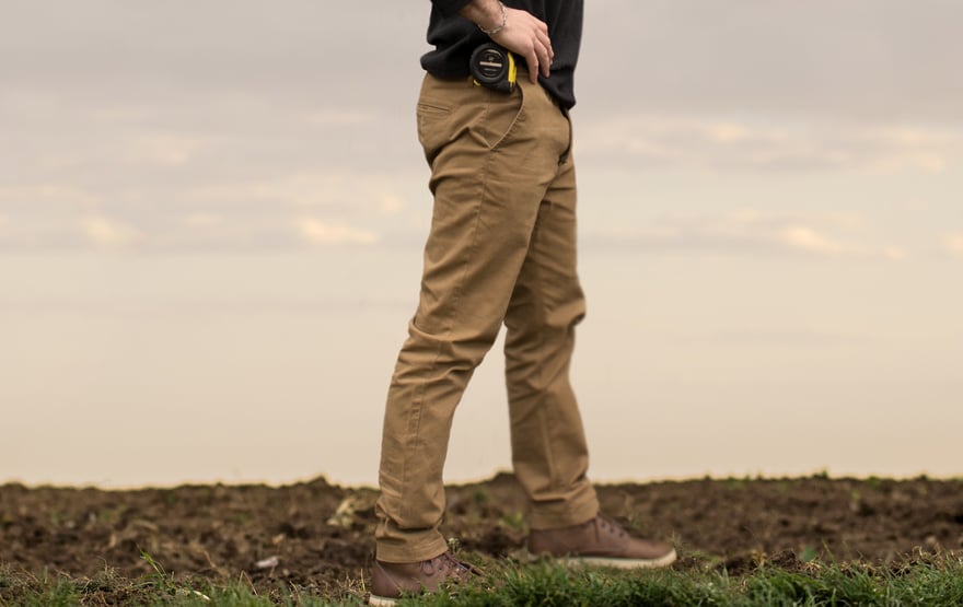 Person wearing tan pants and sneakers, standing in a field. A yellow tape measure is clipped to the front pocket.