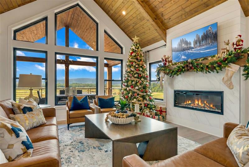 Cozy living room with large windows, a decorated Christmas tree, and a lit fireplace. Warm tones, festive decor, and a scenic view outside.