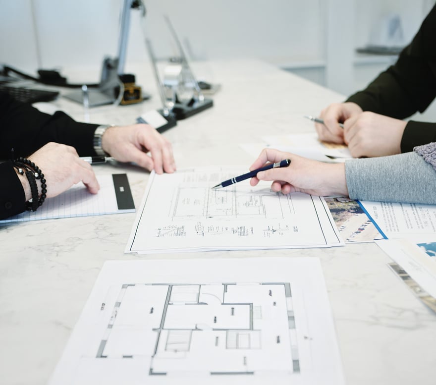 A construction team gathers around a table covered in blueprints, marking plans with a pen. This image represents the preconstruction meeting phase of a design and construction process.