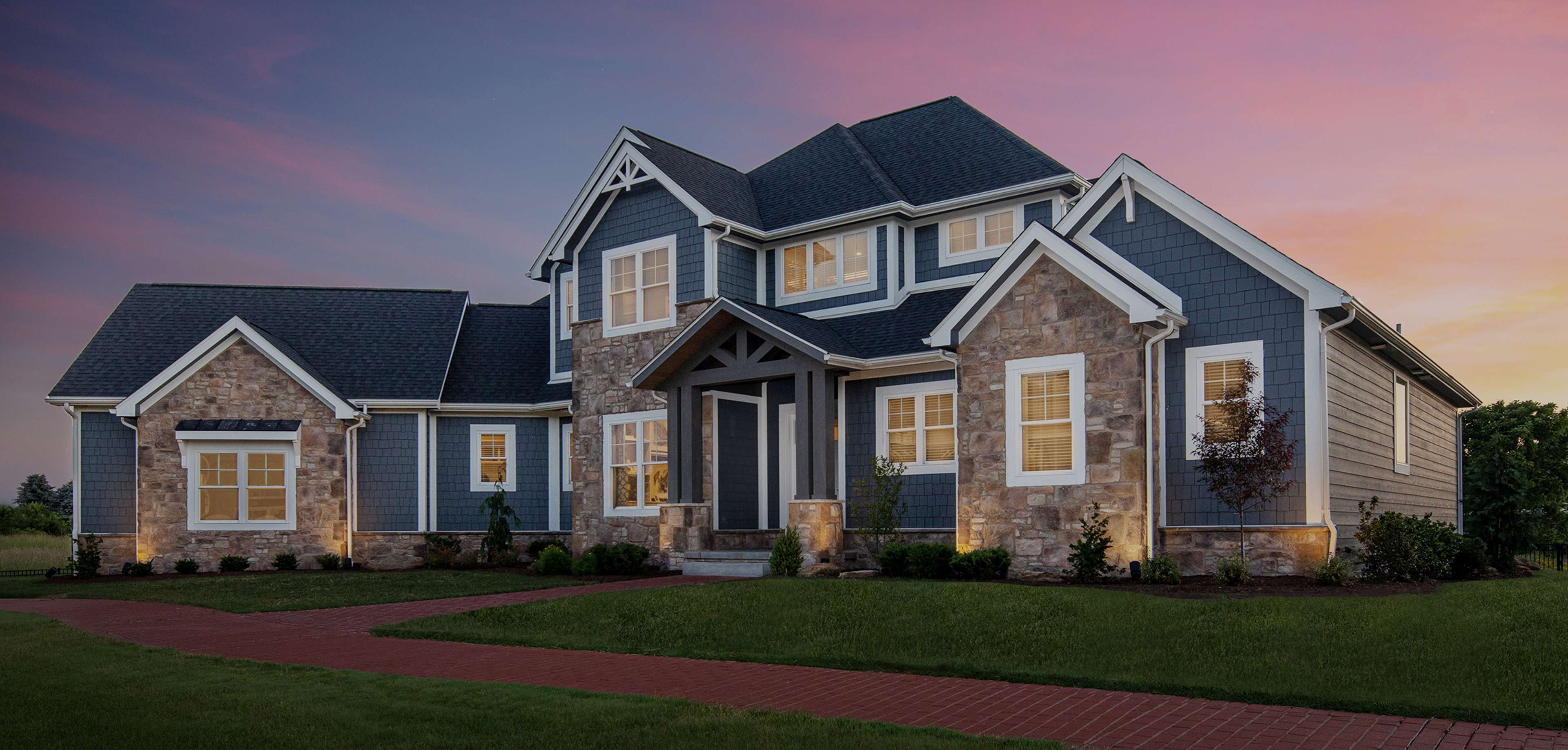 A promotional hero image for a real estate listing, featuring a blue and stone accented luxury home, set against a striking twilight sky. The well manicured lawn and illuminated windows create a welcoming atmosphere.