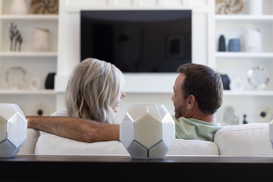 Middle-aged couple facing each other on a couch in a modern home. A tv and decorative vases are displayed behind them on built-in shelving.
