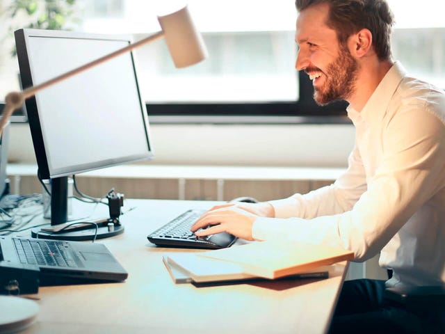Hombre sonriente presentando un examen de alemán en línea desde su computador en una oficina iluminada.