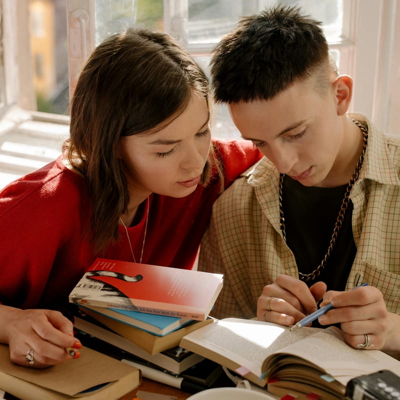 Dos jóvenes sentados leyendo un libro juntos, acompañados de varios libros sobre cuál es el idioma más antiguo del mundo.