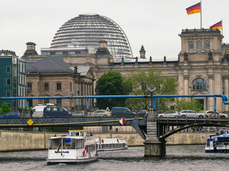 Un edificio con banderas alemanas, un barco en un río y un puente que une las dos orillas.