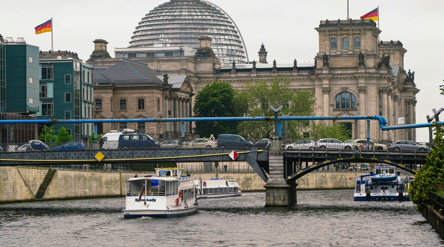 Un edificio con banderas alemanas, un barco en un río y un puente que une las dos orillas.