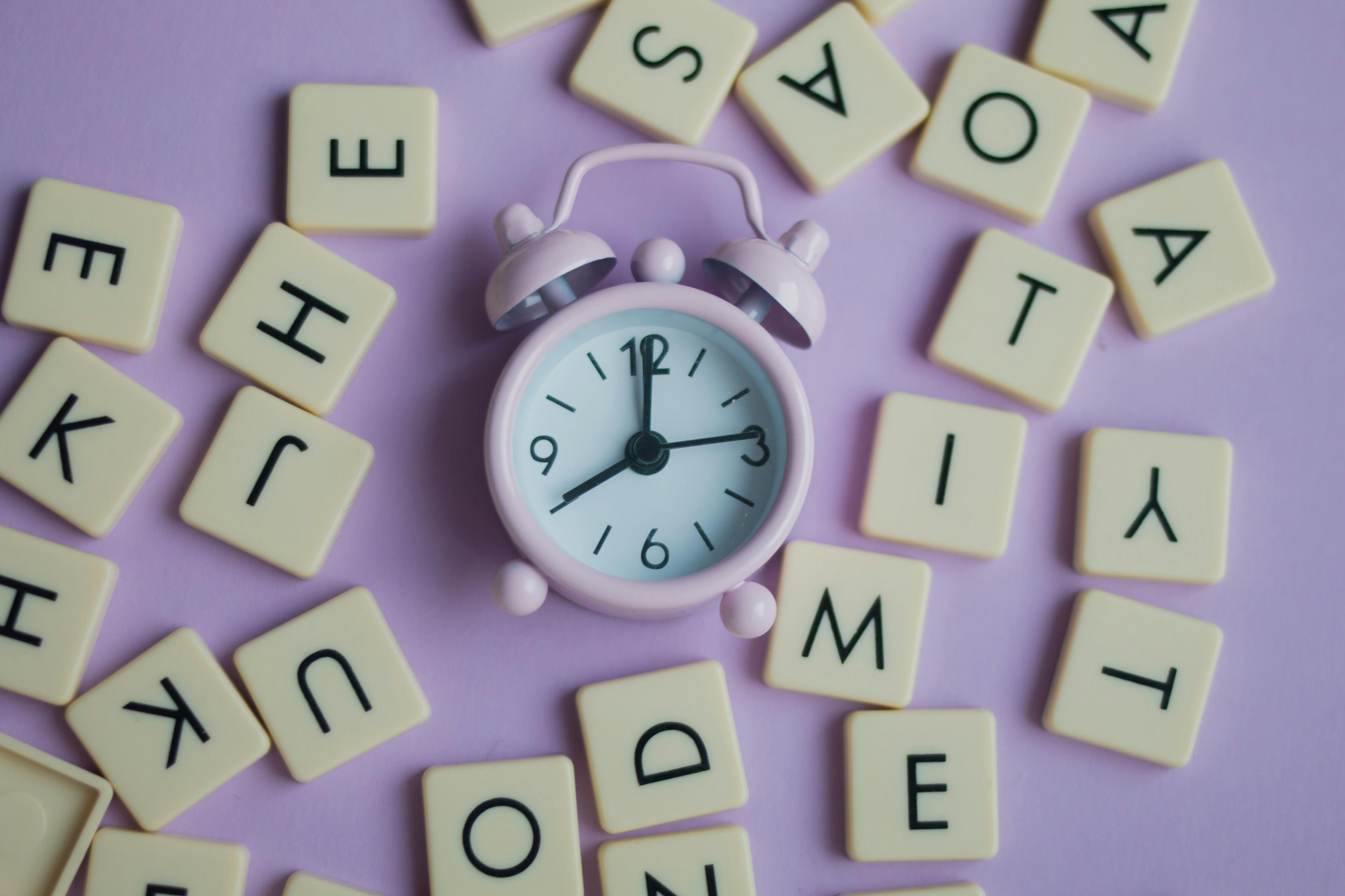 Reloj rosa pastel rodeado de letras en fondo morado pastel para representar la frecuencia en inglés.