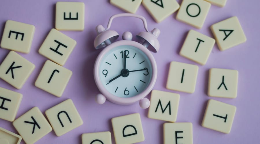 Reloj rosa pastel rodeado de letras en fondo morado pastel para representar la frecuencia en inglés.