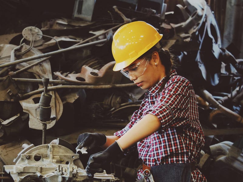 Mujer trabajadora con casco amarillo sosteniendo una pieza mecánica en el Día del Trabajo.