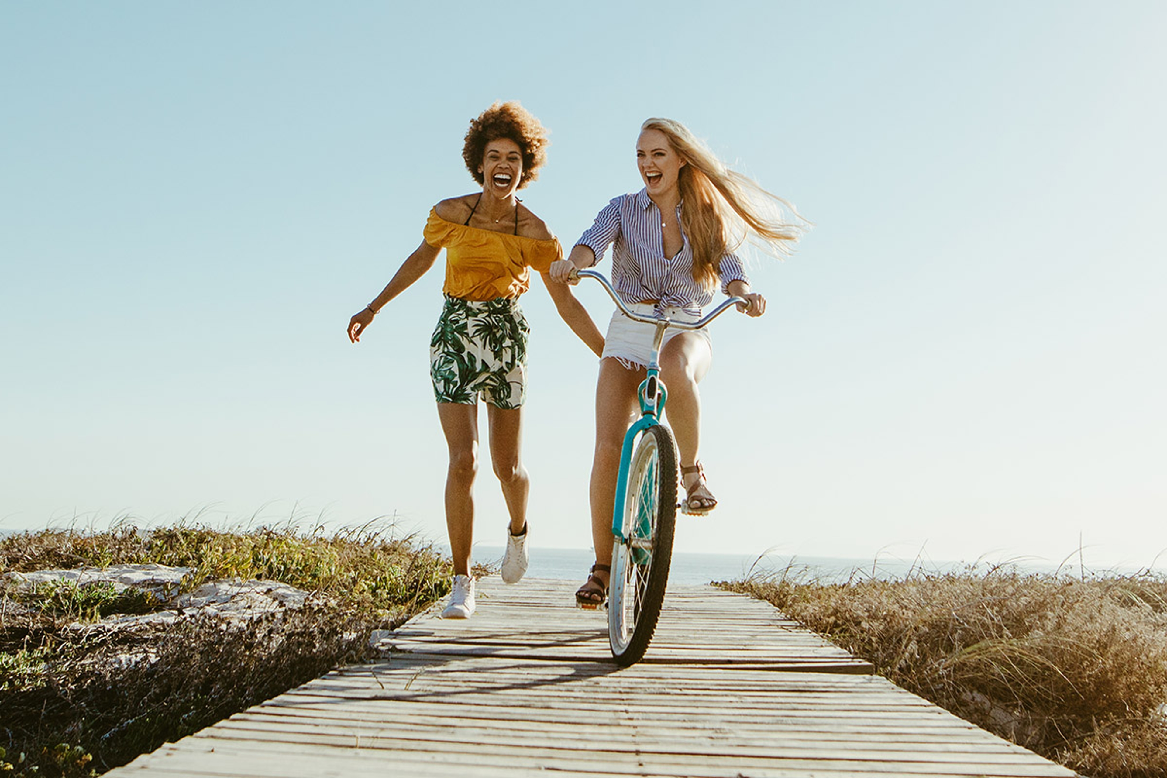 Article Cards Featured Image Excited woman riding bike down the boardwalk with her friends running along. Two female friends having a great time on their vacation. Panoramic shot with lots of copy space on background.