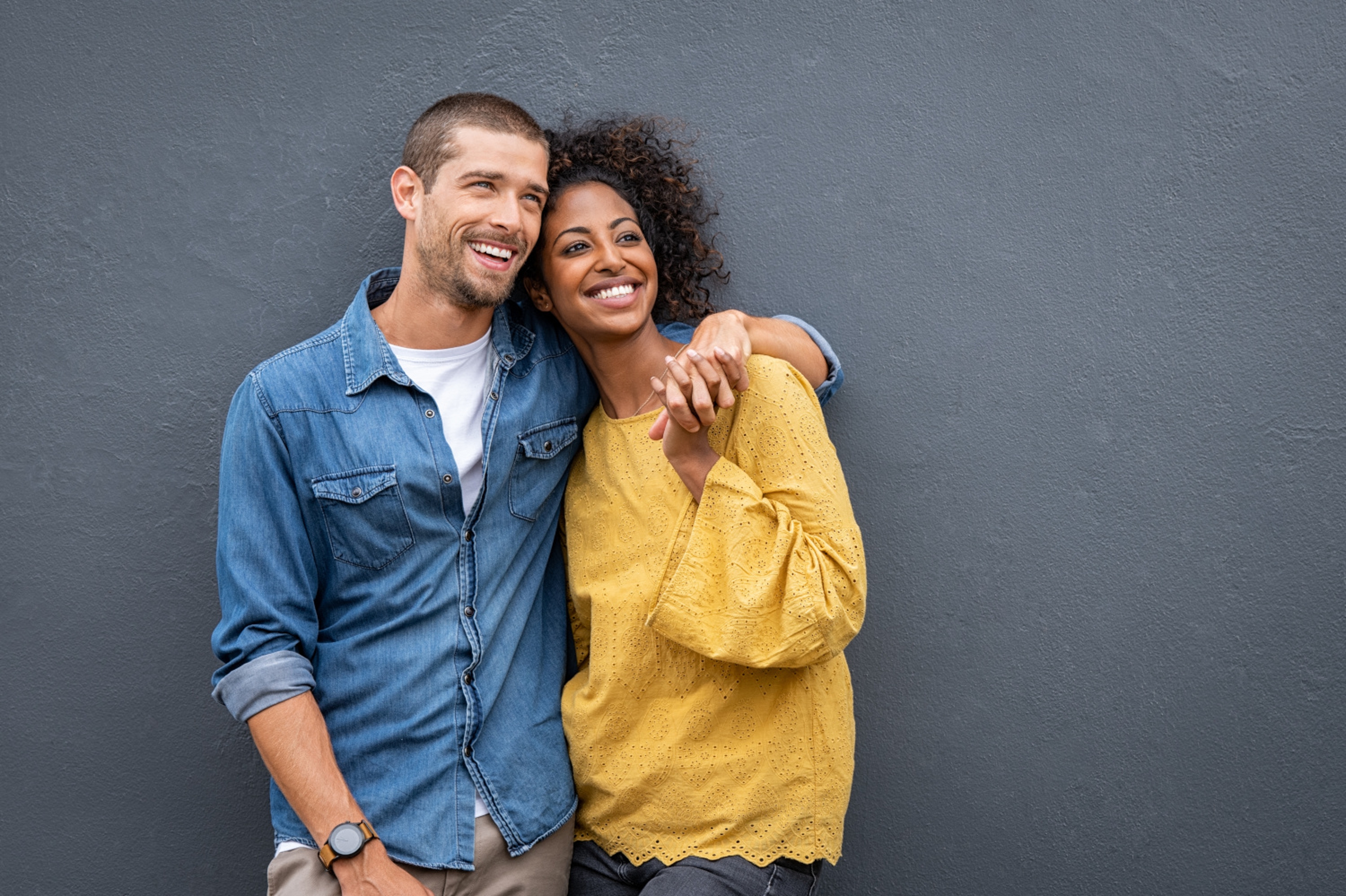 Article Cards Featured Image Young multiethnic couple in love isolated on grey background looking up and thinking about their future together. Smiling man and african woman in casual hugging and looking away while planning the future. Cheerful couple holding hands while leaning on wa