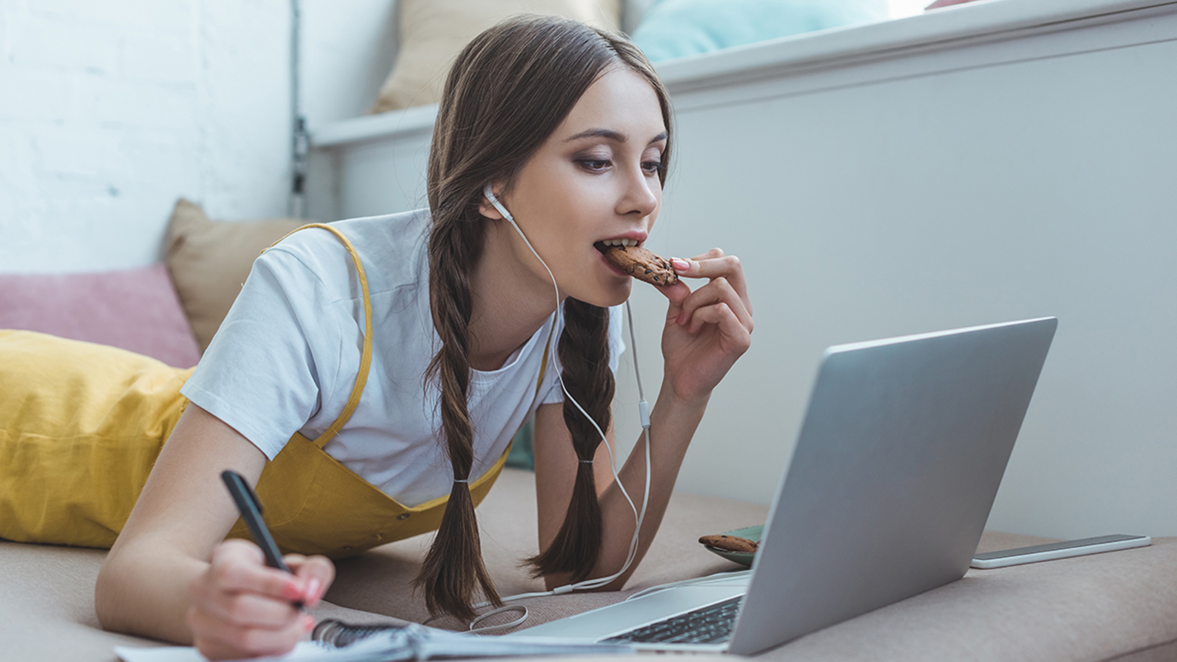 beautiful teen girl eating cookie writing in copybook and using laptop on sofa