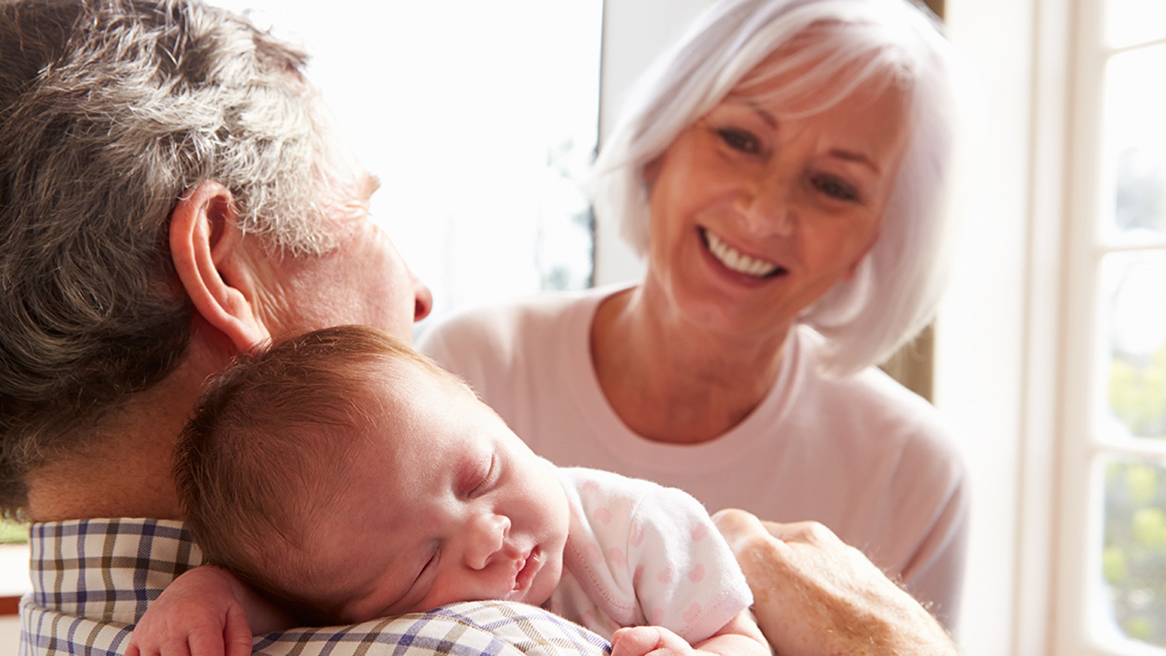 Grandfather holding sleeping grandchild while grandmother smiles at them.