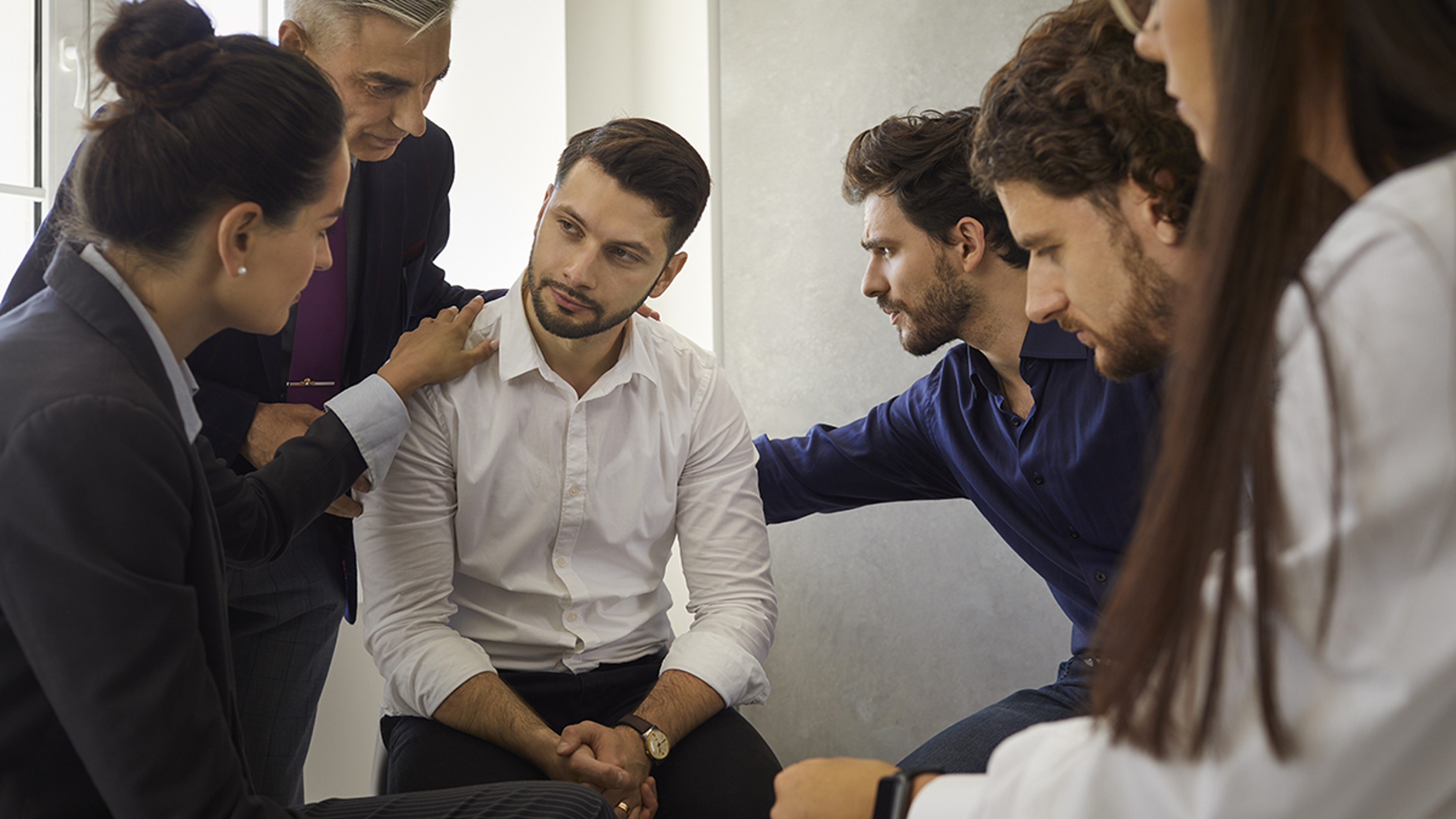 Article Cards Featured Image Group of different people sitting in a circle supporting and comforting a sad upset young man. Concept of psychological help, group therapy and overcoming problems together