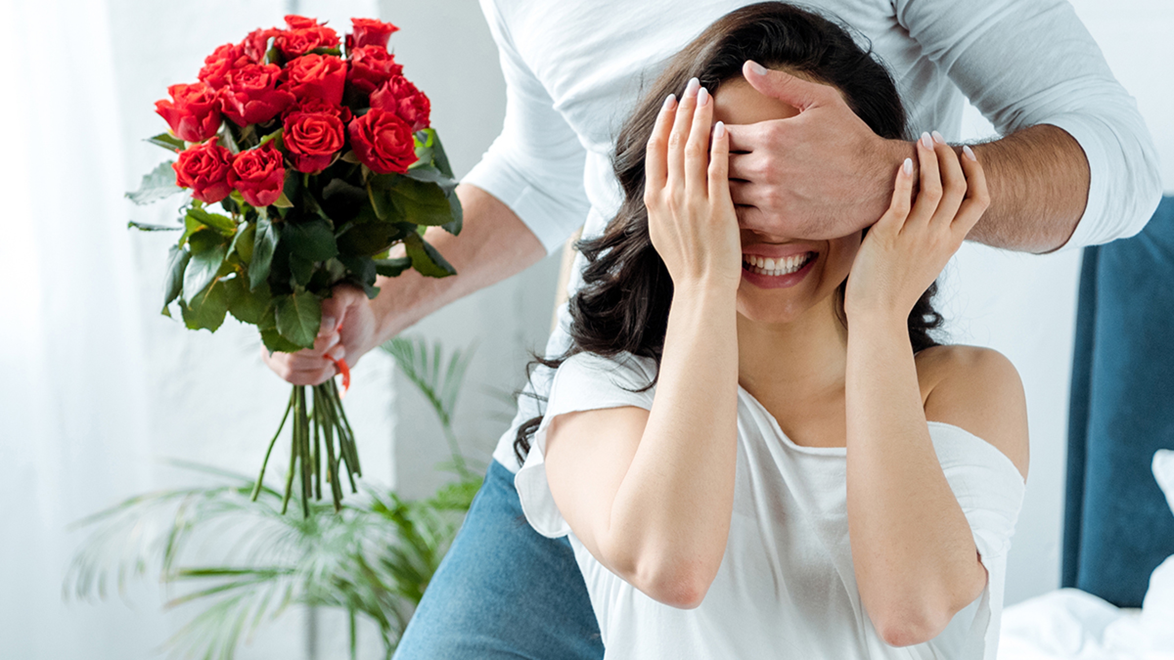 partial view of a man covering a womens eyes to surprise her with a bouquet of roses