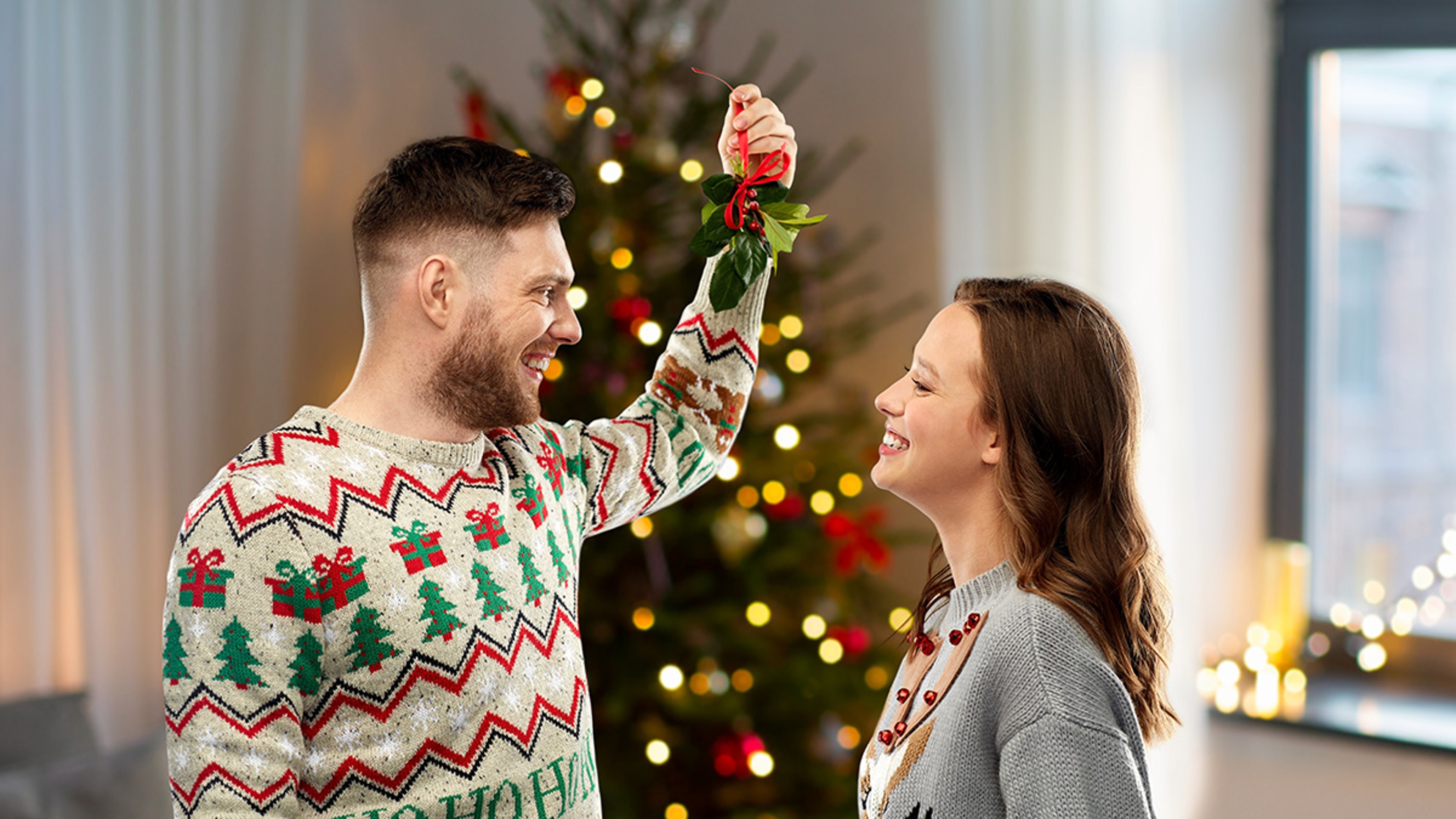 christmas, people and holiday traditions concept  portrait of happy couple in ugly sweaters with mistletoe over home background