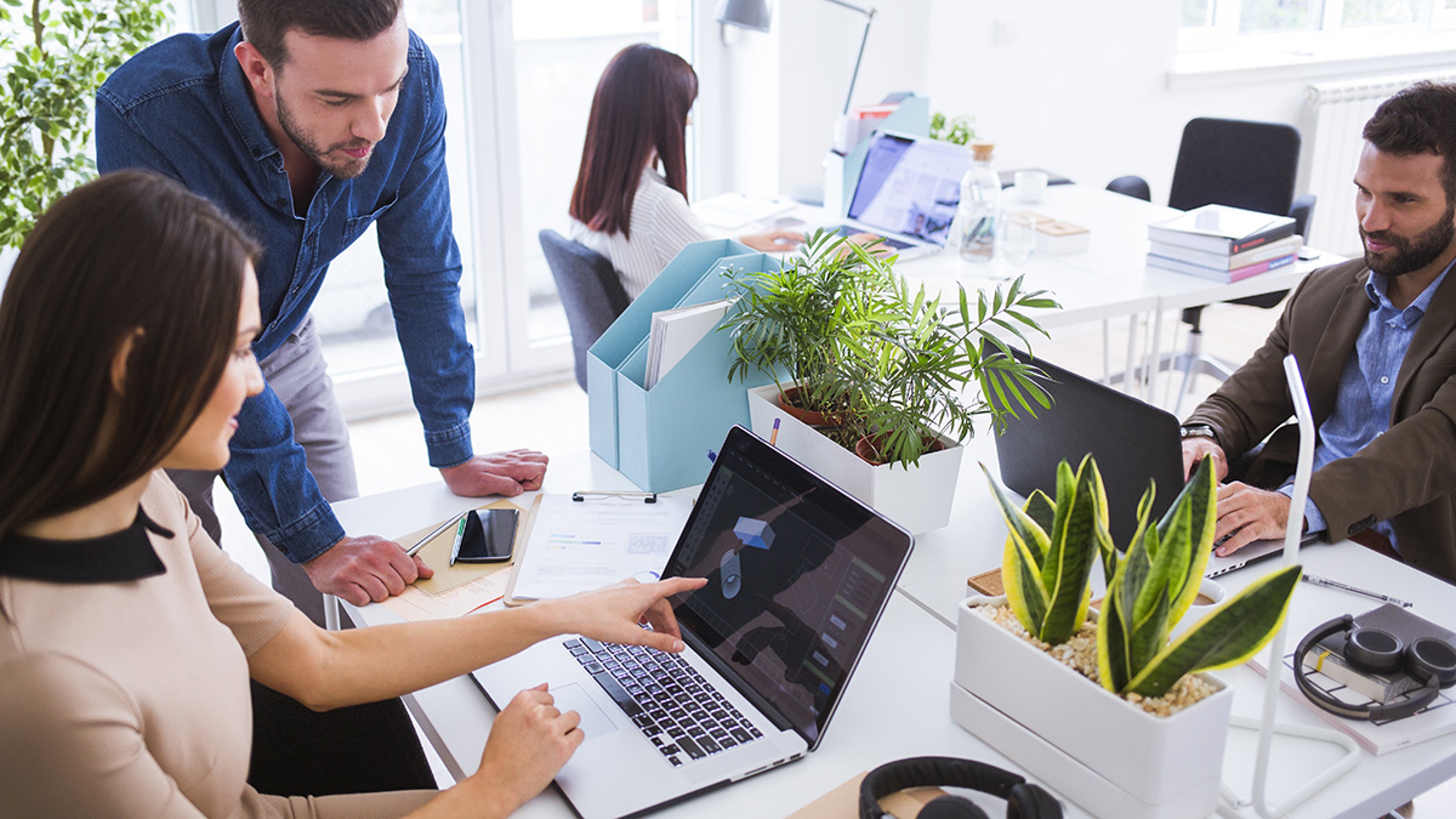 Group of young people in smart casual wear communicating and using modern technologies while working in an office surrounded by green desk plants