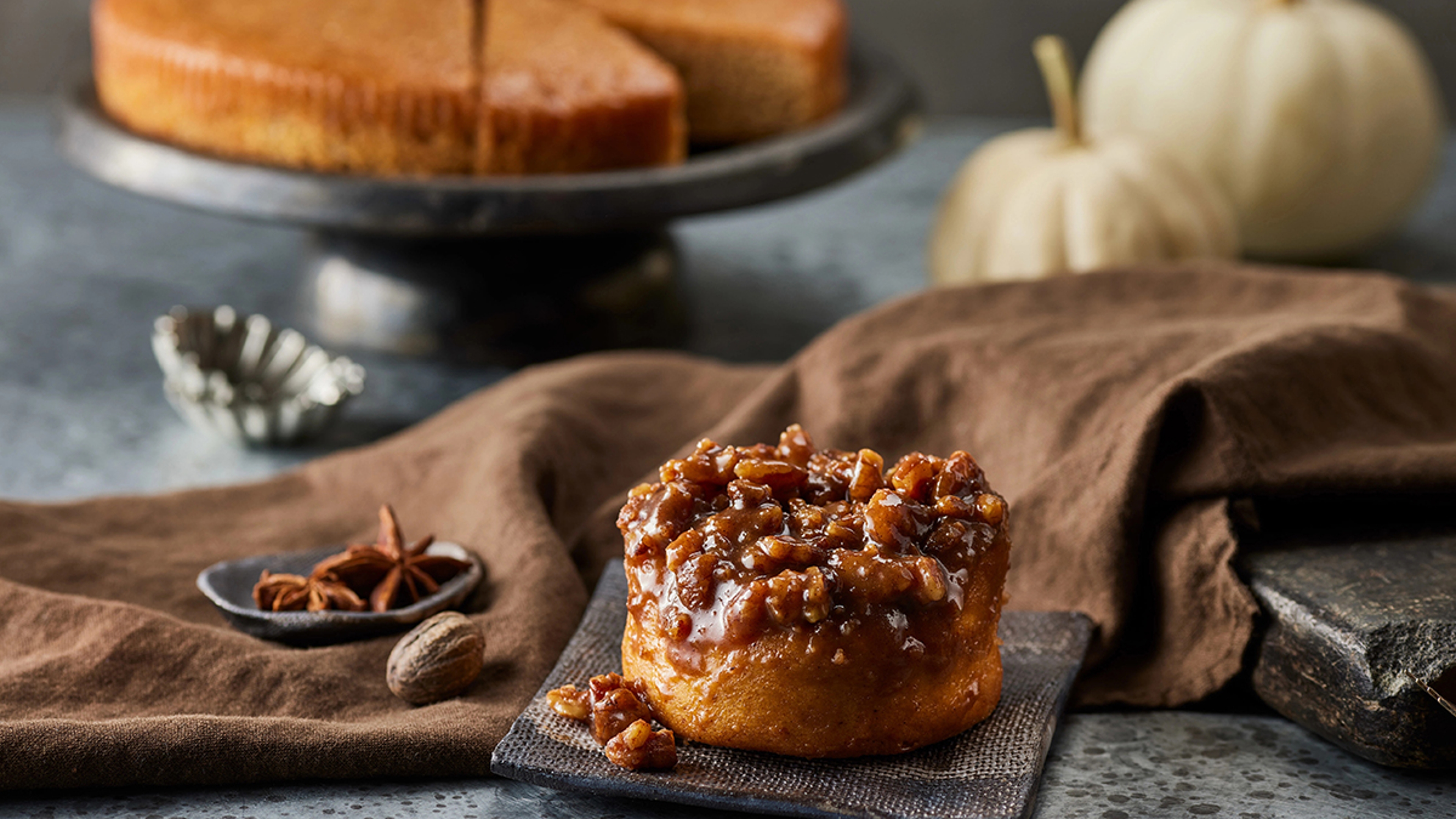 Cinnamon roll topped with nuts with cake and pumpkins in the background.