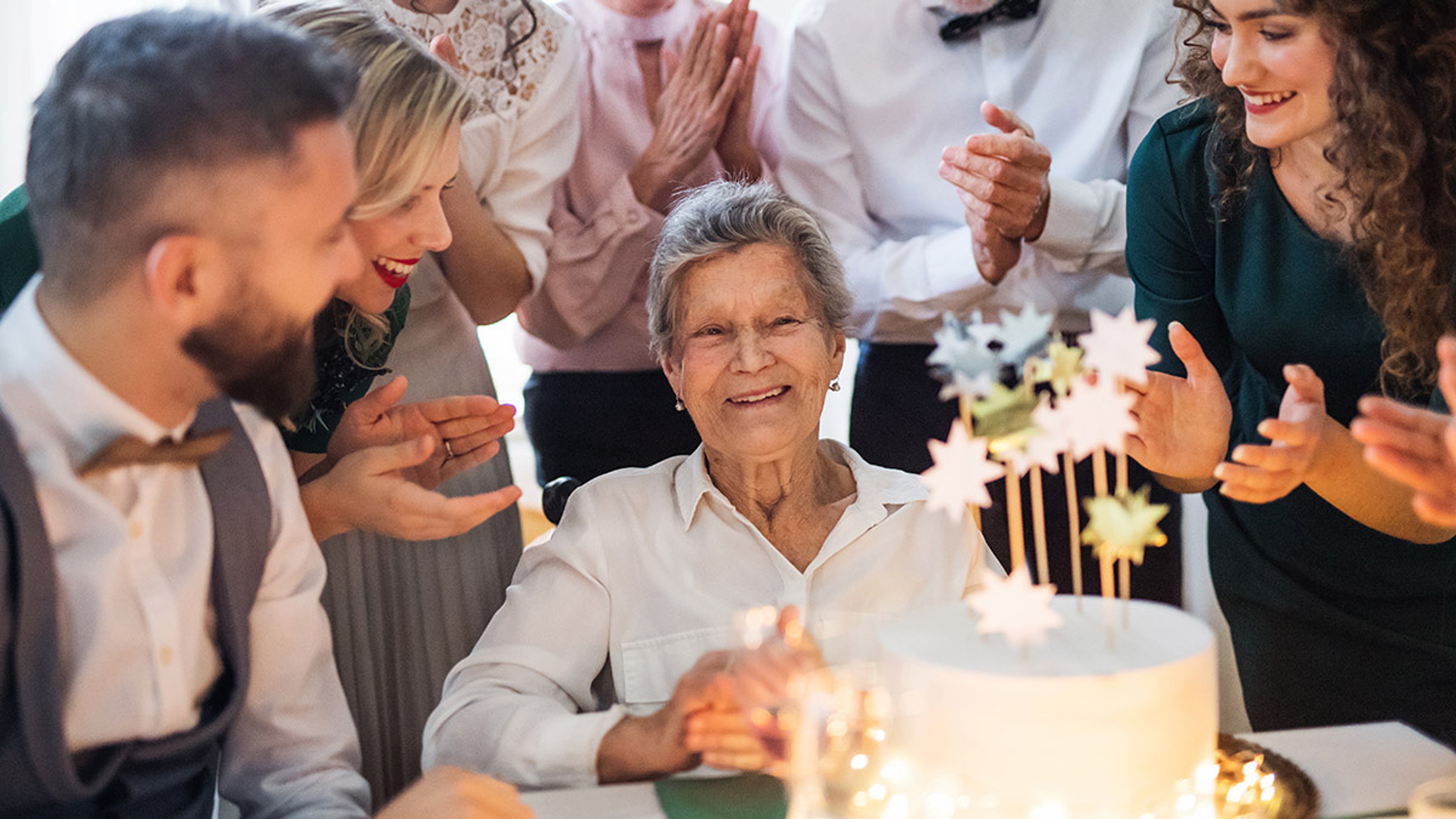 Older woman smiling at a cake surrounded by family.