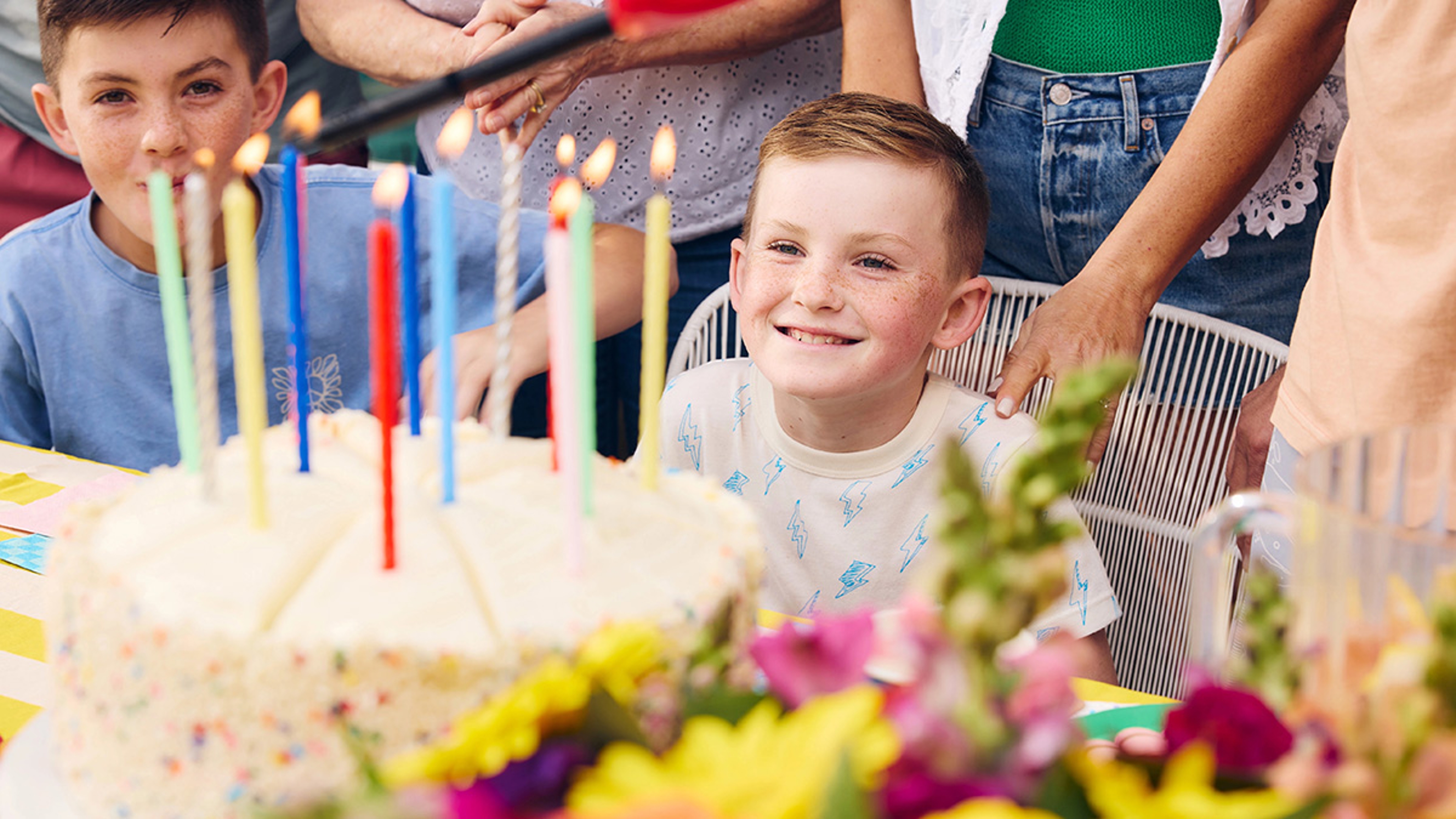 Boy smiling at birthday cake while surrounded by people singing happy birthday.