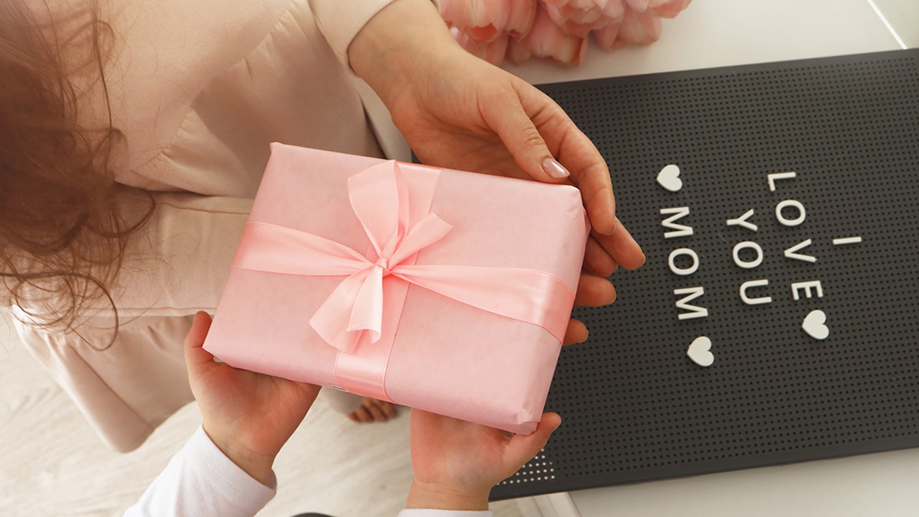 Child's hands hold beautiful pink gift box with ribbon. Pink tulip flower and sign i love you mom in background. Top view, close up. Preparing for the holidays
