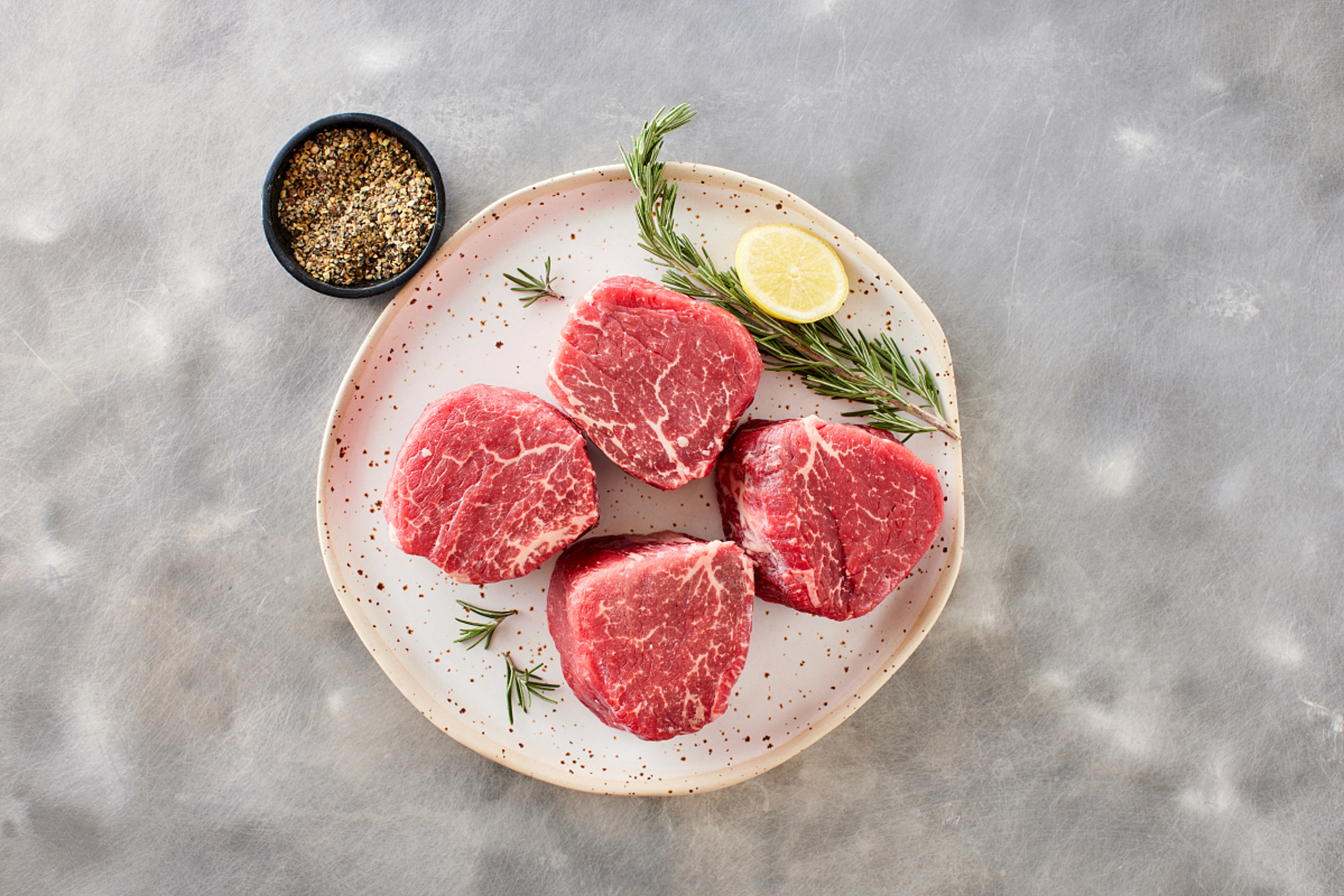 Four filet mignon cuts on a plate with lemon and rosemary next to a bowl of seasoning.