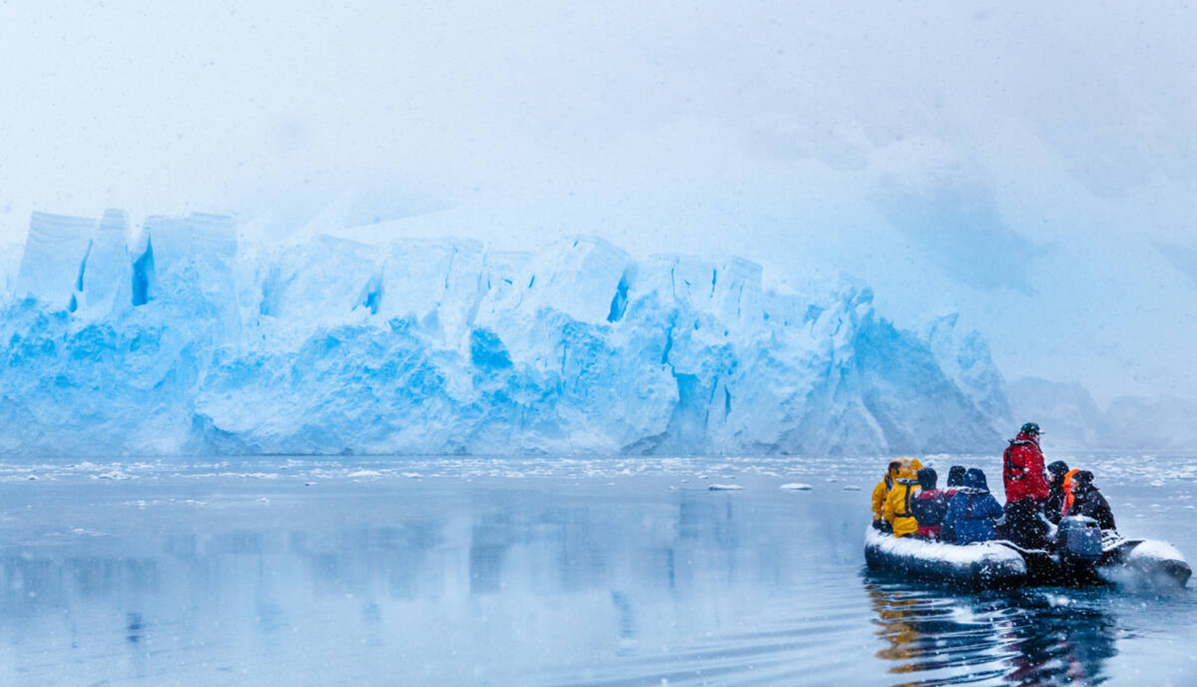 Snowfall over the boat with frozen tourists driving towards the huge blue glacier wall in the background, near Almirante Brown, Antarctic peninsula Snowfall over the boat with frozen tourists driving towards the huge blue glacier wall in the background, near Almirante Brown, Antarctic peninsula