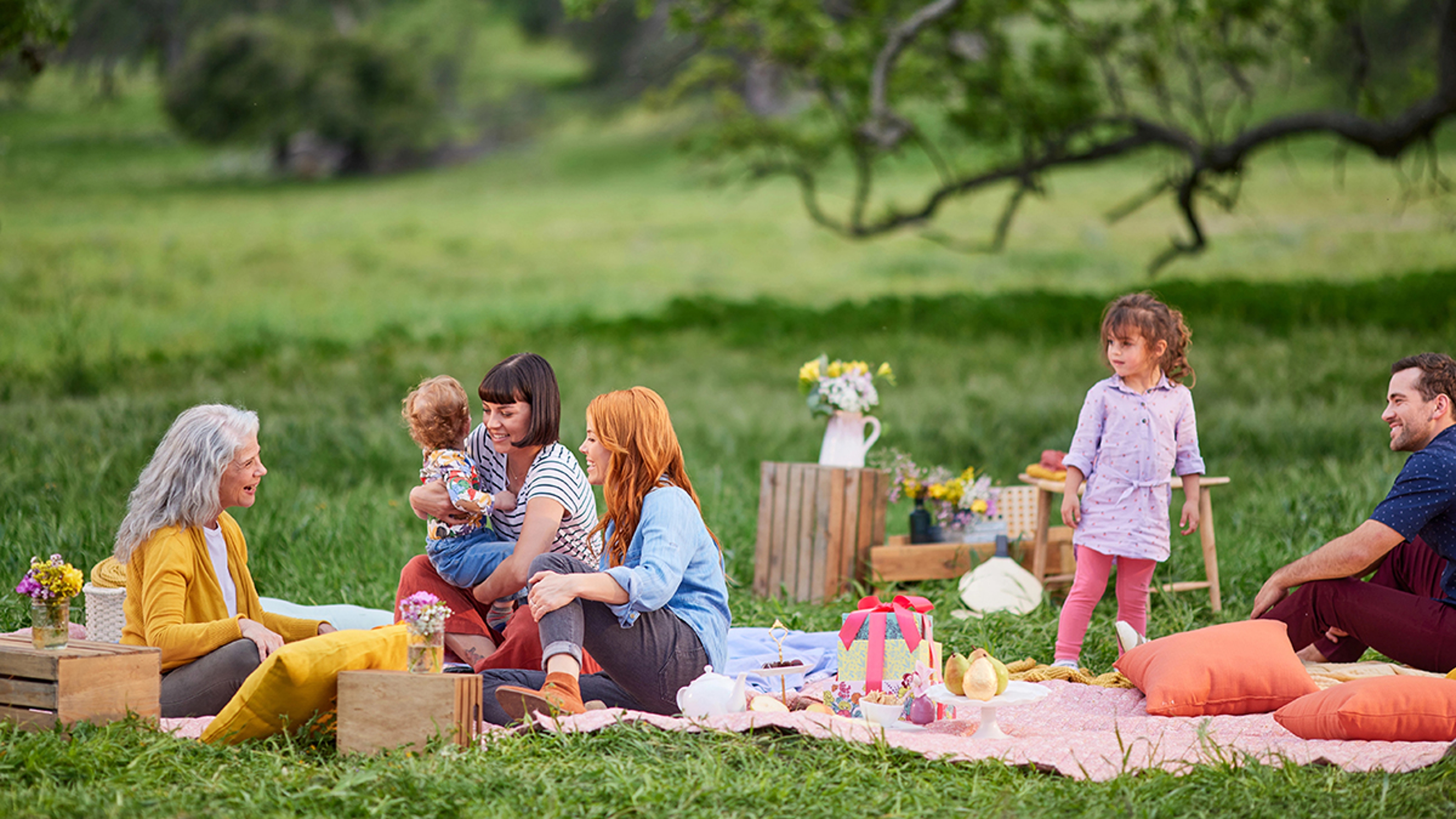 A photo of picnic with a group of people sitting outside on a blanket surrounded by food and gifts