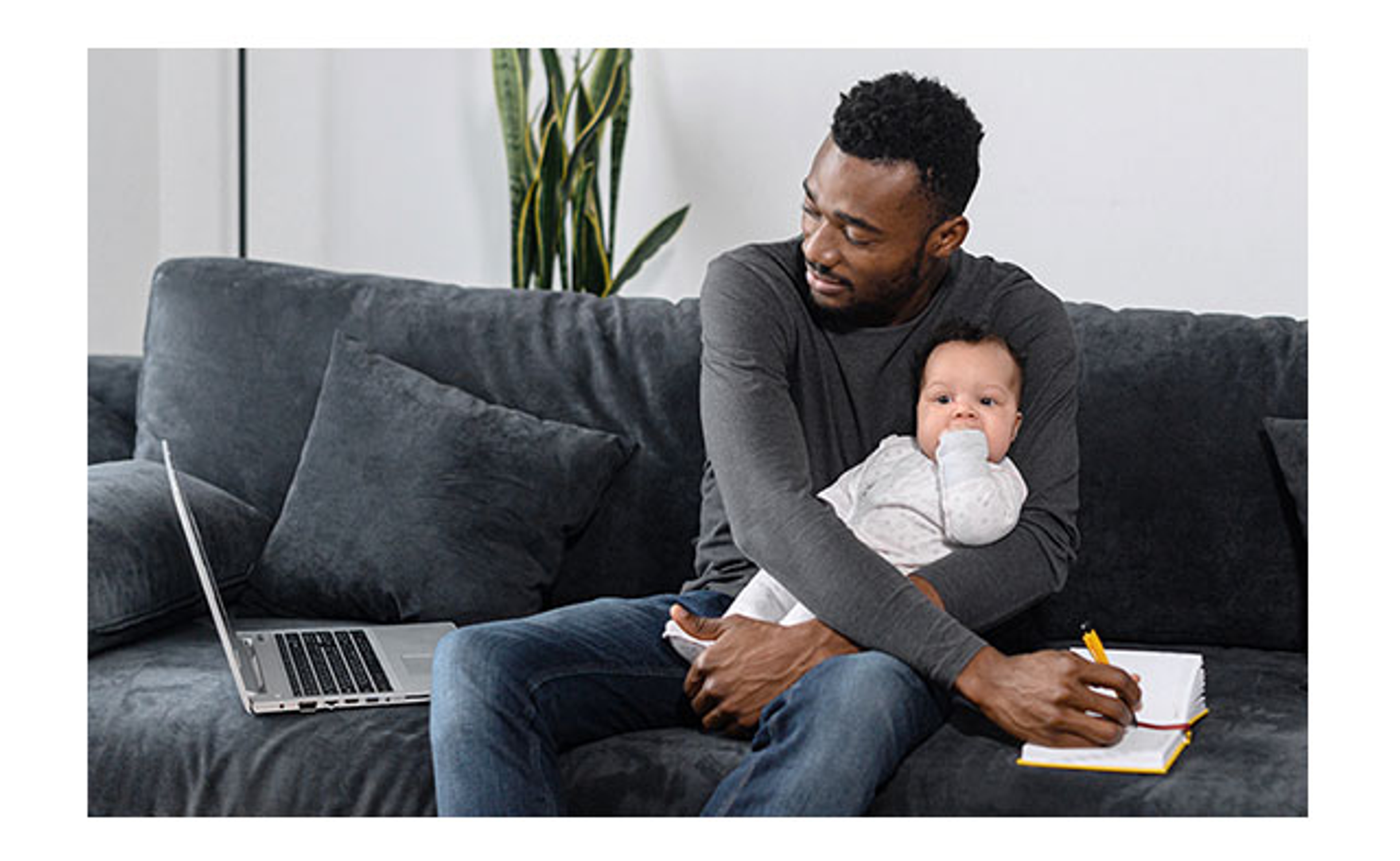 Dad on a couch working on a laptop while cradling his baby