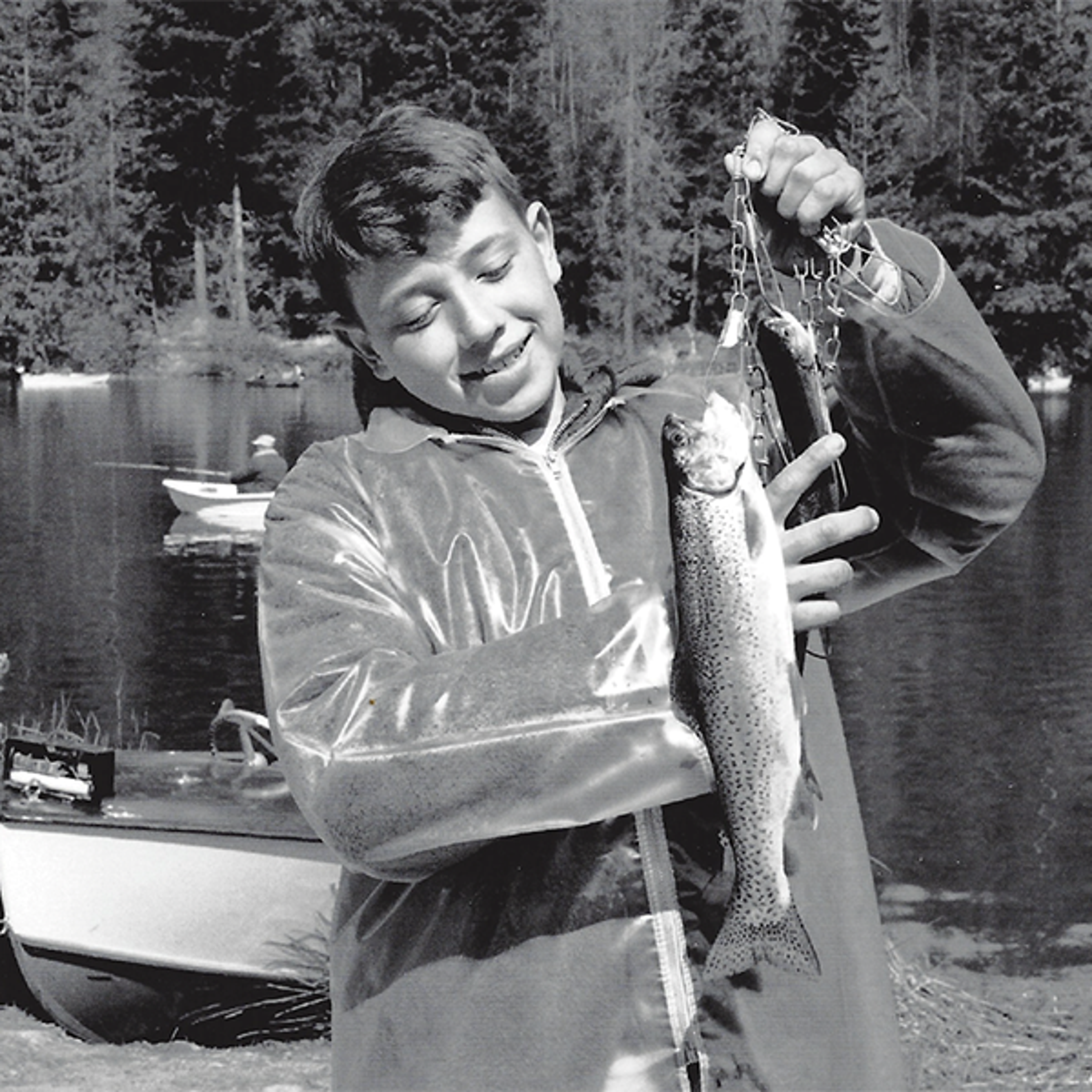 A photo of vital choice with a young boy holding up a fish on a line that's caught in the lake behind him.