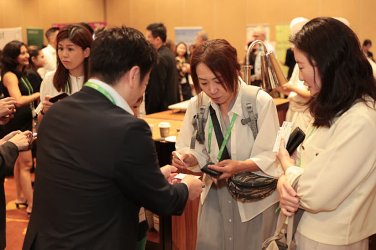 Attendees seated at round tables during a conference session, taking notes and listening to speakers. Water pitchers and notebooks are visible on tables.