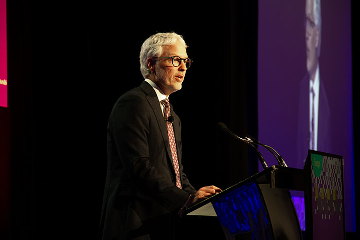 Speaker at a conference podium delivering a presentation, standing beside a microphone on a stage with dark curtains and a large projected screen in the background.
