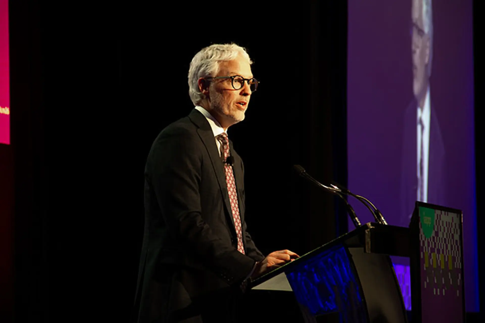 Speaker at a conference podium delivering a presentation, standing beside a microphone on a stage with dark curtains and a large projected screen in the background.