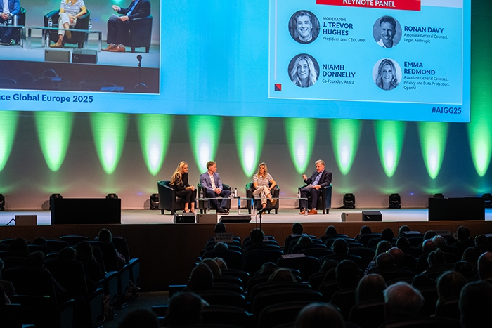 Panel discussion on a brightly lit stage with four speakers seated in black chairs. A large screen behind displays names and photos of the keynote panel.