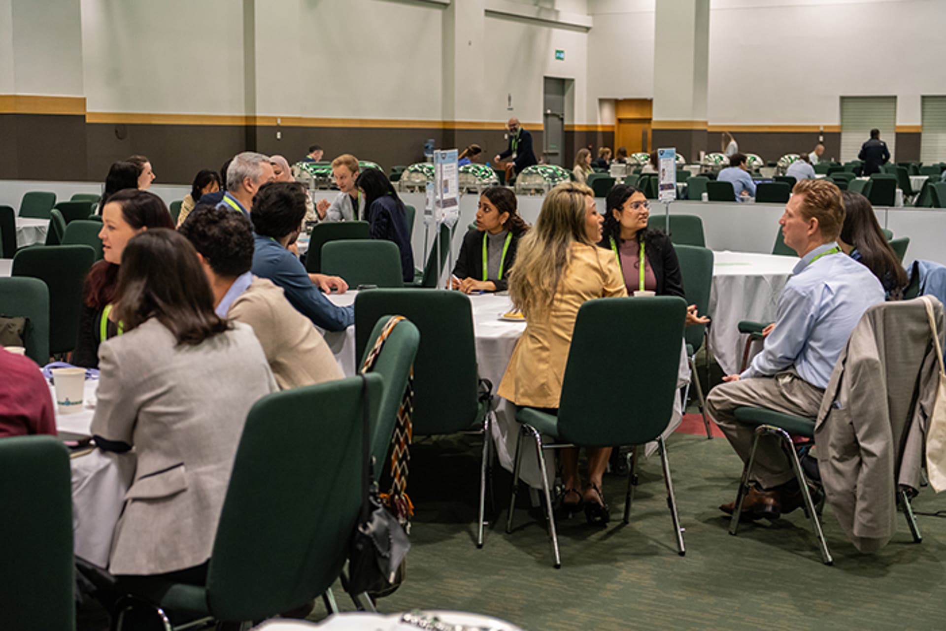 Multiple small groups seated at round tables in a large conference room, engaged in discussions. Informational signs are placed on tables.