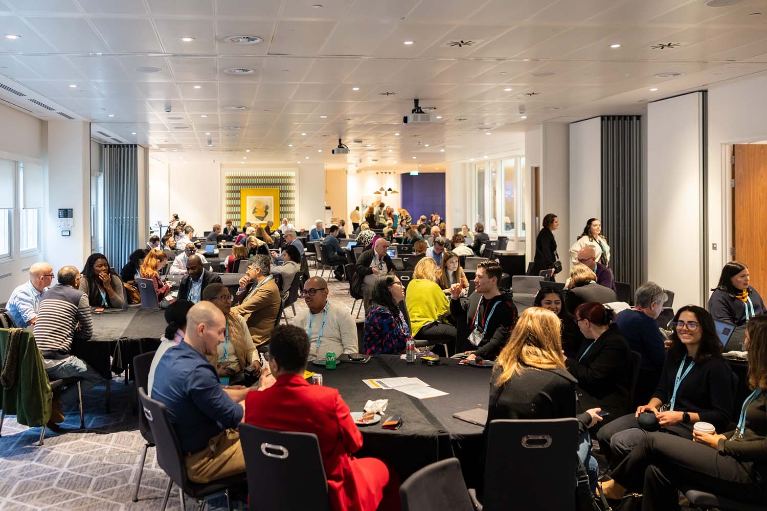 Participants sit at round tables engaged in group discussions during a conference session in a large meeting room. The space is well lit with attendees spread across multiple tables.