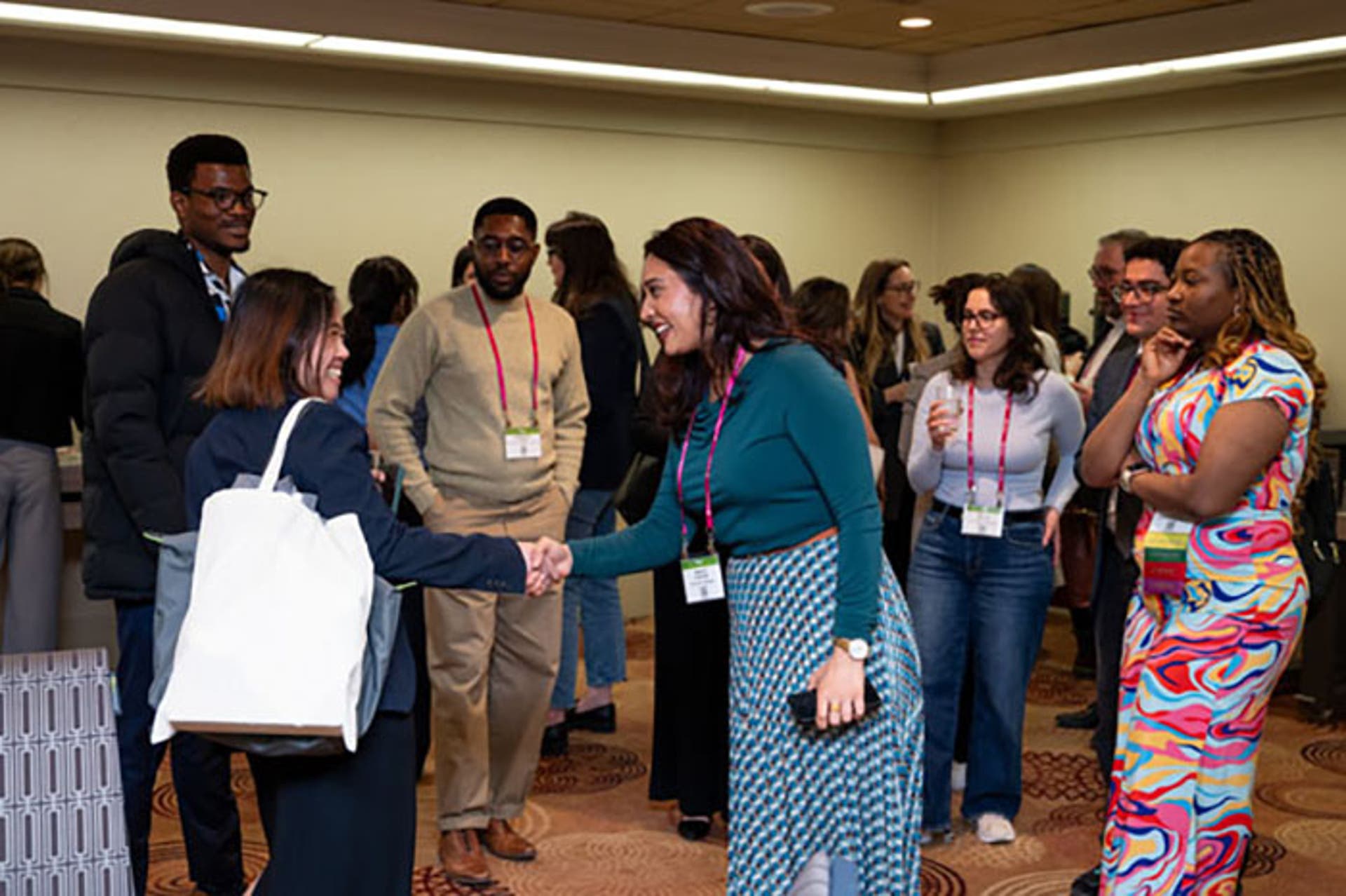 Two attendees shaking hands in a meeting room during a networking session, while others stand and talk nearby.