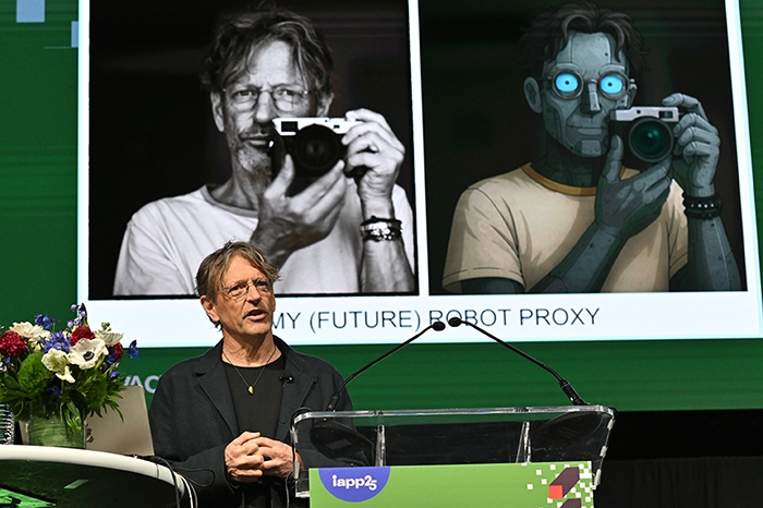 Speaker standing at a podium with a large screen behind showing two side-by-side images: one of a person holding a camera and another of a robot holding a camera. Text on the screen reads “MY (FUTURE) ROBOT PROXY.” A floral arrangement is on the podium.
