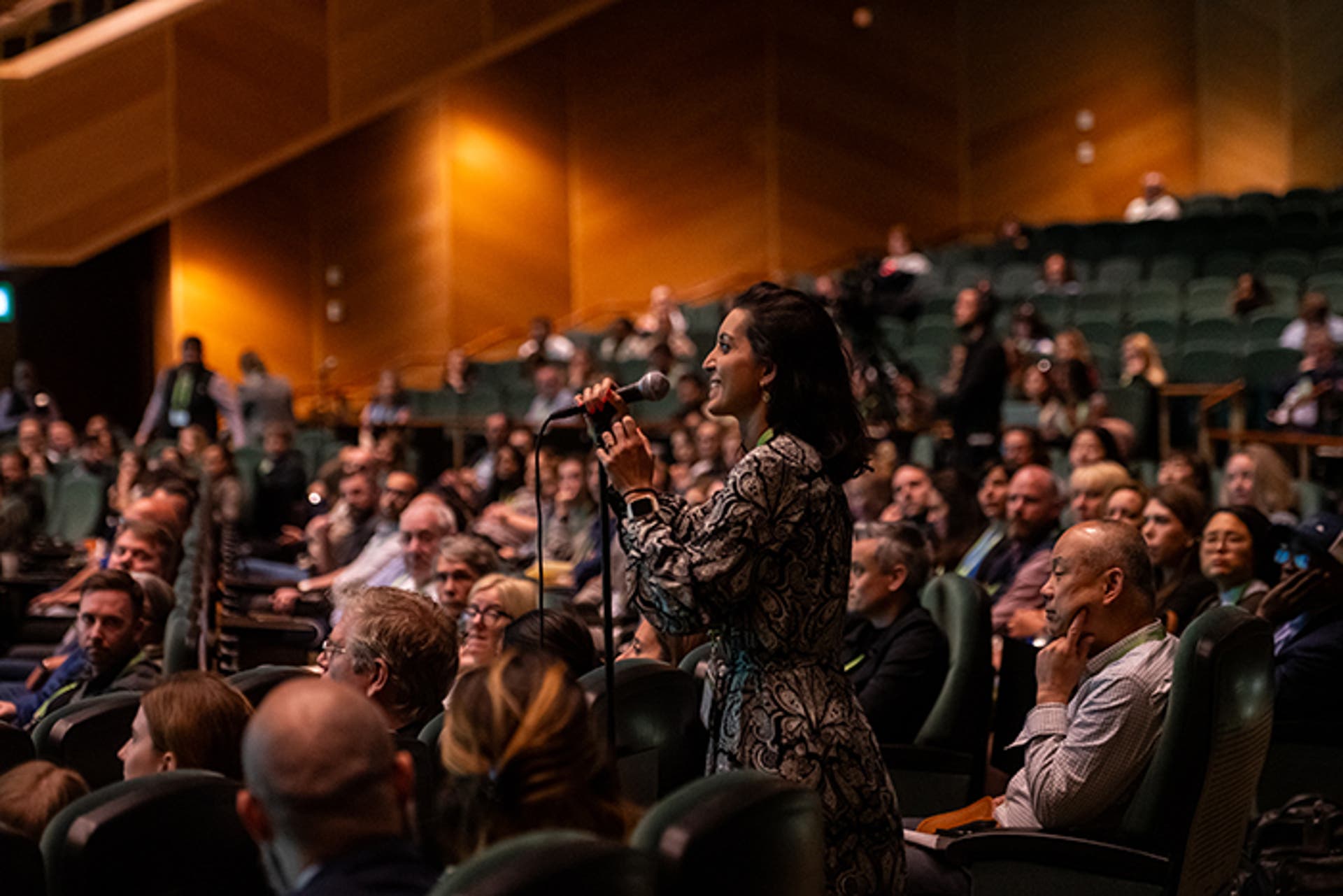 Attendee standing at a microphone in an auditorium during a Q&A session. Audience members are seated in rows behind.