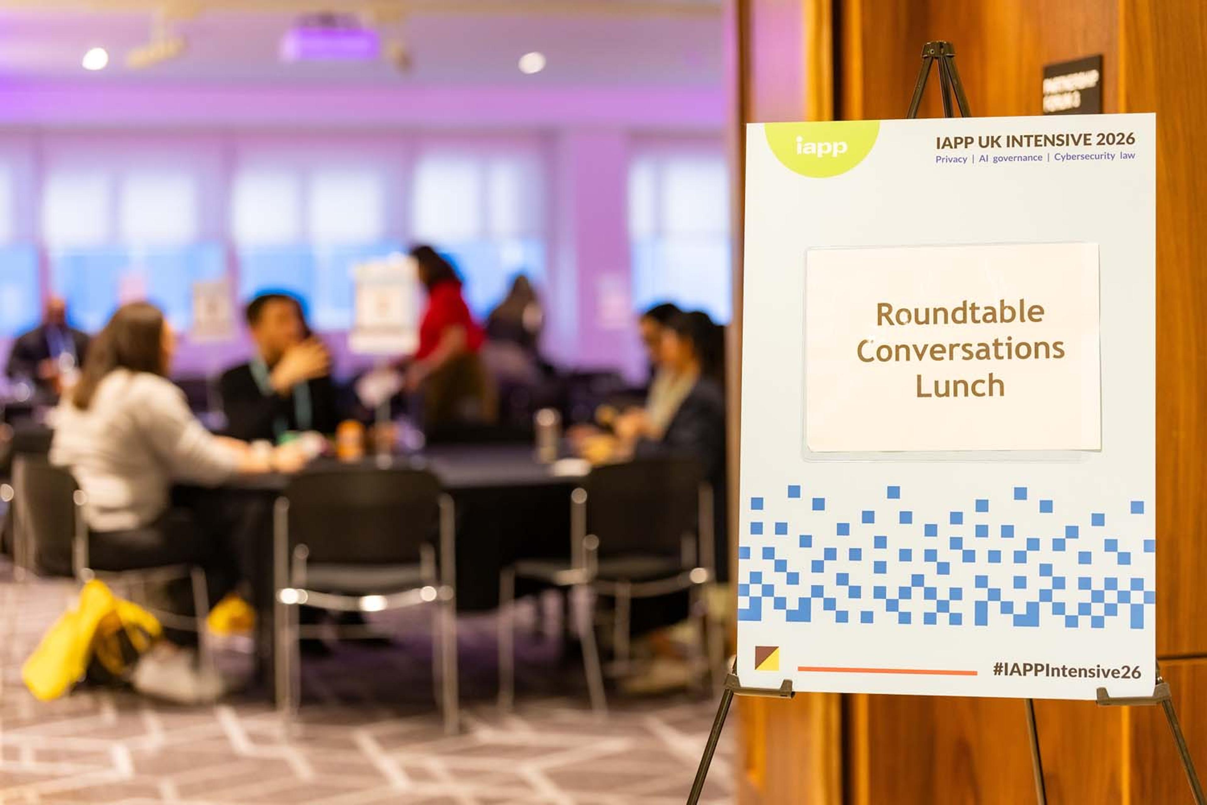 A sign on an easel reads Roundtable Conversations Lunch for IAPP UK Intensive 2026 in the foreground. Conference attendees sit at round tables in the background participating in small group discussions.