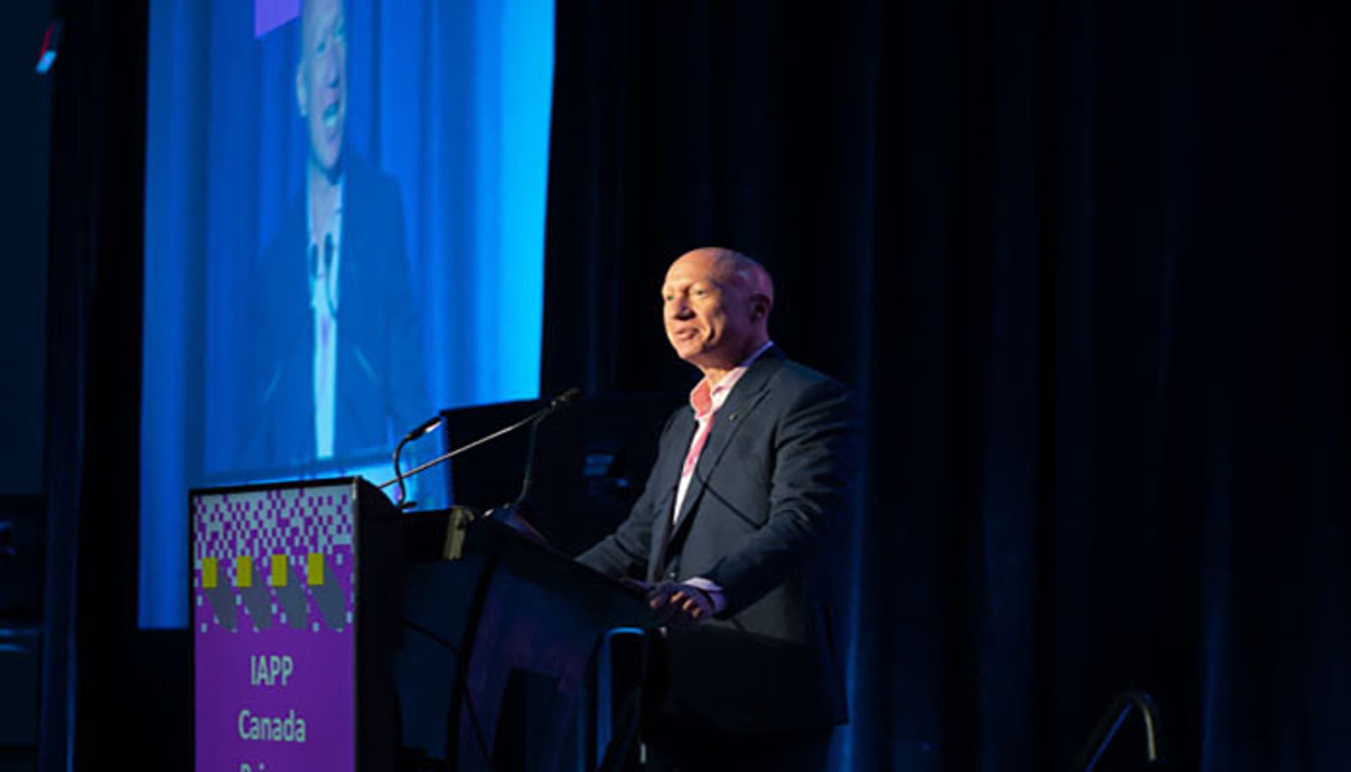 Speaker at a podium on a stage with a blue curtain backdrop. A large pink sign on the podium reads “IAPP Canada Privacy Symposium 2025,” and the speaker’s image is projected on a screen.