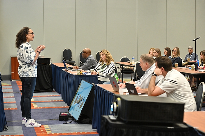 Presenter standing and speaking to a group seated at tables in a conference room. A large monitor on the floor displays a slide, and laptops are visible on the tables.