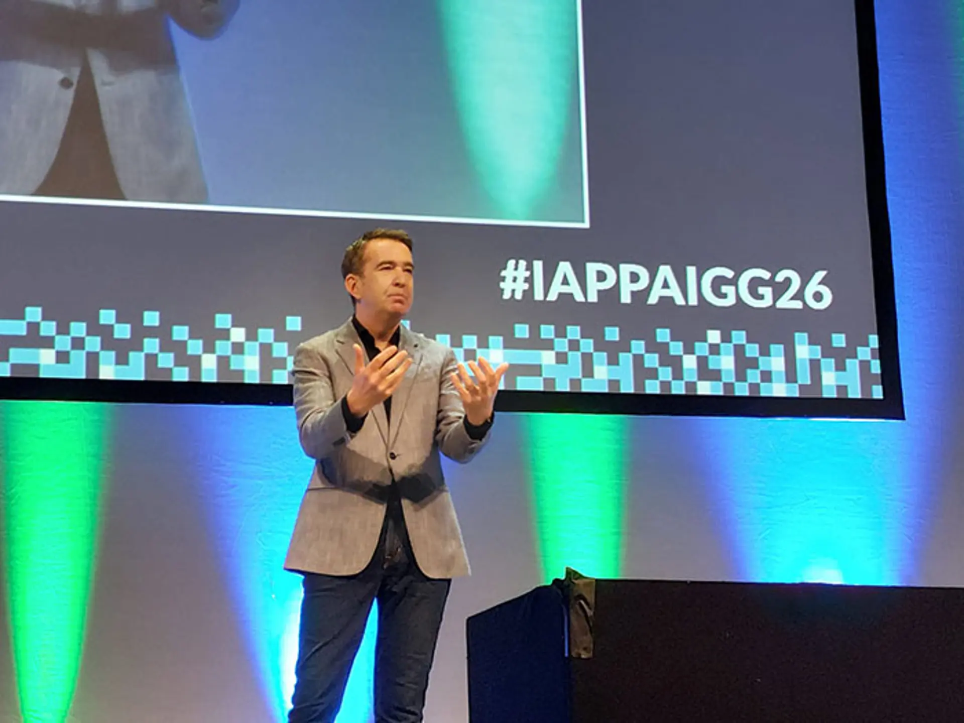 A presenter stands on a stage in front of a large screen displaying the hashtag IAPPAIGG26 and gestures with both hands while addressing an audience. The stage lighting features blue and green tones, and a podium and table are visible nearby on the stage.