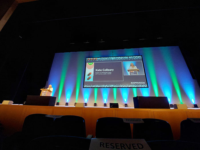 A conference stage is viewed from the audience with several empty seats in the foreground labeled reserved. A speaker stands at a podium on the left side while a large screen displays a presentation slide with the name Kate Colleary and the event hashtag IAPPGAC26, illuminated by blue and green stage lighting.
