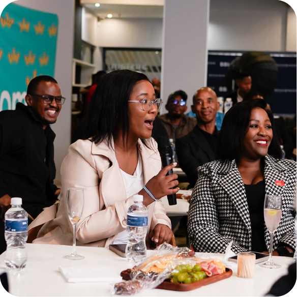 A seated person holding a microphone at a table with drinks and a charcuterie board while others sit and stand around in an indoor event space.