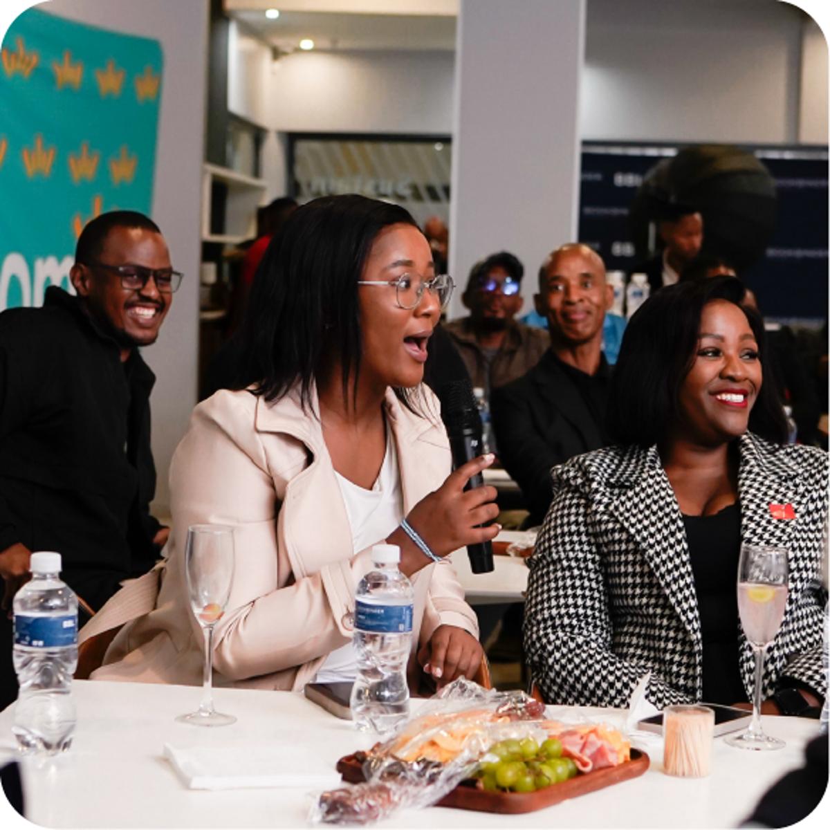 A seated person holding a microphone at a table with drinks and a charcuterie board while others sit and stand around in an indoor event space.