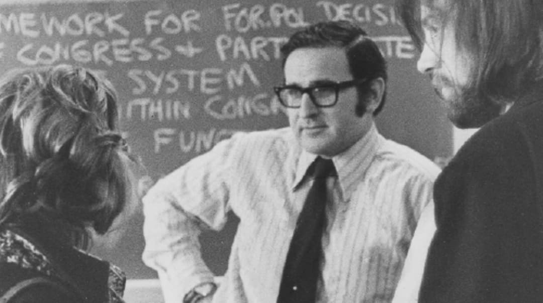 Dr. Alan Westin stands with students; he is in glasses and a tie with his hand on his hip. Behind him is a chalkboard covered in notes.