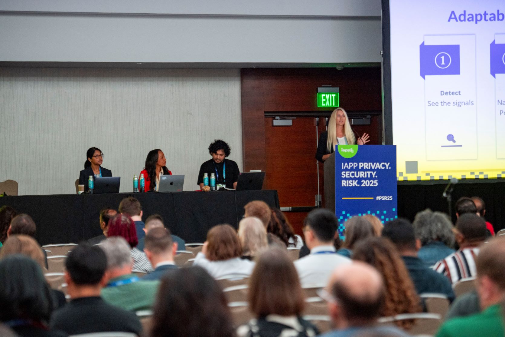 Conference session with a speaker standing at a podium labeled “IAPP Privacy. Security. Risk. 2025” in front of a large screen displaying a slide titled “Adaptability” with bullet points including “Detect: See the signals.” Three panelists are seated at a long table with laptops and water bottles. Audience members are seated in rows facing the stage.